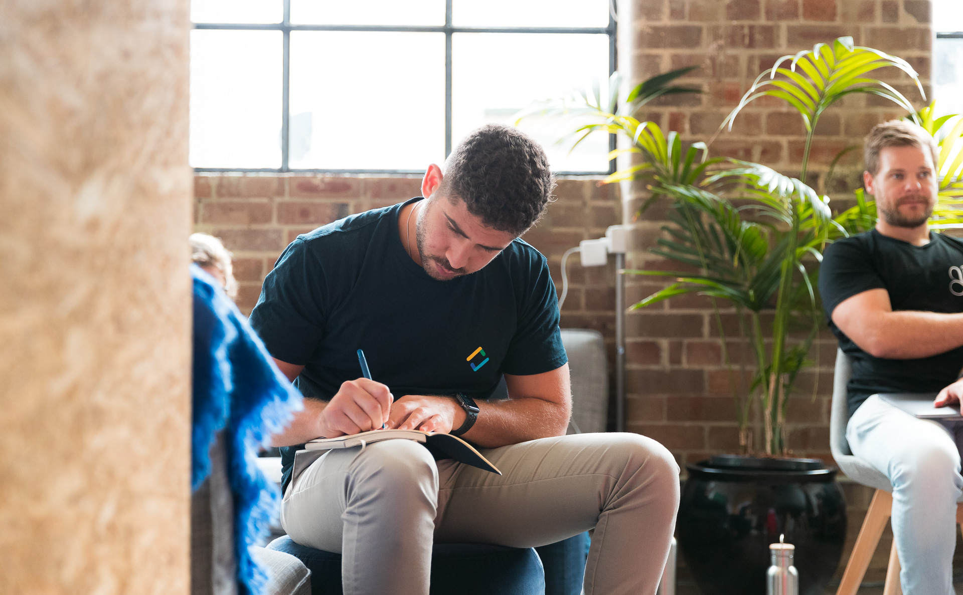 Person in dark shirt writing in notebook in modern brick office space with plants and large windows.